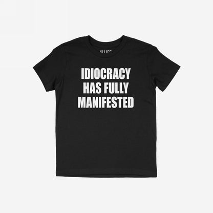 Black crew-neck T-shirt featuring the slogan IDIOCRACY HAS FULLY MANIFESTED in bold white letters, conveying a satirical and politically charged message.