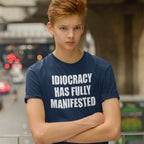 Teenage boy on an overpass wears the Idiocracy T-shirt, featuring bold white text, IDIOCRACY HAS FULLY MANIFESTED, reflecting a blunt, politically disillusioned statement amid blurred city traffic below.