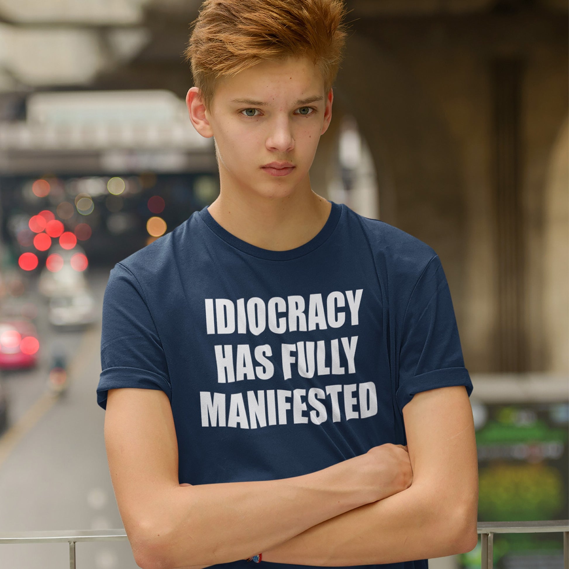 Teenage boy on an overpass wears the Idiocracy T-shirt, featuring bold white text, IDIOCRACY HAS FULLY MANIFESTED, reflecting a blunt, politically disillusioned statement amid blurred city traffic below.