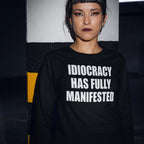 Woman in industrial space leans on a striped pillar, wearing the Idiocracy T-shirt with bold white text, “IDIOCRACY HAS FULLY MANIFESTED,” capturing a blend of style and defiant expression.