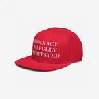 Red flat-brimmed snapback hat with white serif text IDIOCRACY HAS FULLY MANIFESTED embroidered on the front. Reflects bold social commentary, aligning with themes of equality and anti-fascism.