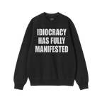 Black crewneck sweatshirt with bold white text reading IDIOCRACY HAS FULLY MANIFESTED, reflecting a political statement. Bran