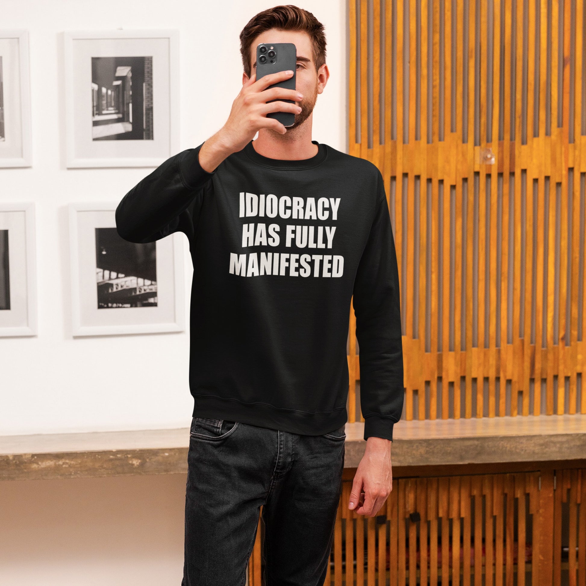 Person taking a selfie wearing the Idiocracy Sweatshirt, featuring bold white text “IDIOCRACY HAS FULLY MANIFESTED,” paired with dark jeans, set in a minimalist interior with framed photos and wooden panels.