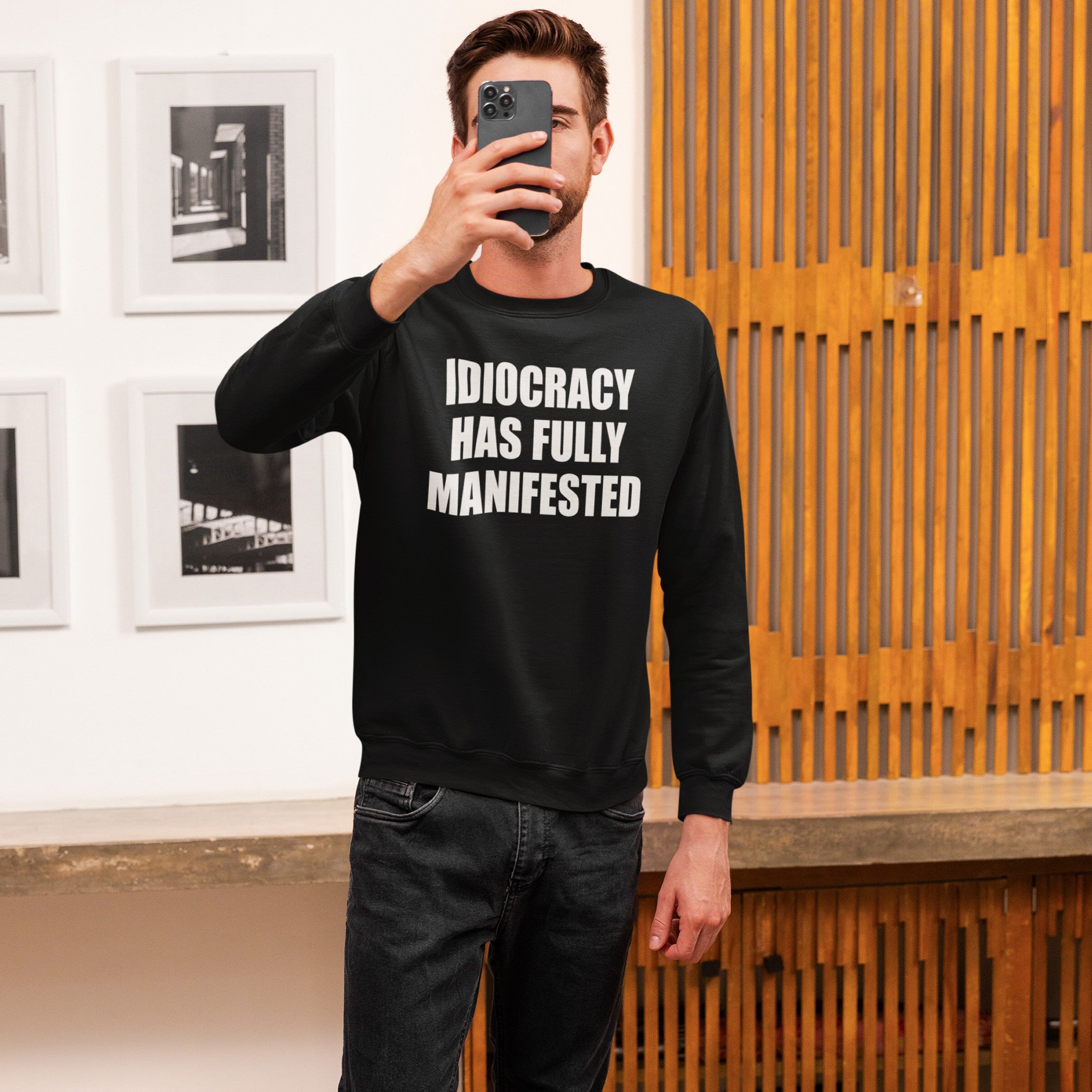 Person taking a selfie wearing the Idiocracy Sweatshirt, featuring bold white text “IDIOCRACY HAS FULLY MANIFESTED,” paired with dark jeans, set in a minimalist interior with framed photos and wooden panels.
