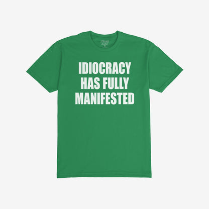 Green, short-sleeved Idiocracy T-shirt features bold white text stating “IDIOCRACY HAS FULLY MANIFESTED”, reflecting a satirical take on current events, ideal for the politically disillusioned and meme enthusiasts.
