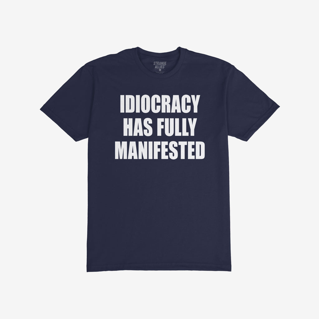 The Idiocracy T-shirt showcases bold white text stating IDIOCRACY HAS FULLY MANIFESTED on a dark-navy background, reflecting a sharp social commentary for the politically disillusioned.