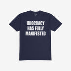 The Idiocracy T-shirt showcases bold white text stating IDIOCRACY HAS FULLY MANIFESTED on a dark-navy background, reflecting a sharp social commentary for the politically disillusioned.