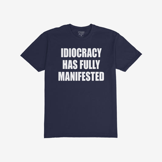 The Idiocracy T-shirt showcases bold white text stating IDIOCRACY HAS FULLY MANIFESTED on a dark-navy background, reflecting a sharp social commentary for the politically disillusioned.