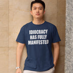 Young man wearing the Idiocracy T-shirt with bold white text, “IDIOCRACY HAS FULLY MANIFESTED,” standing against a tiled wall, embodies a statement piece for the politically aware.