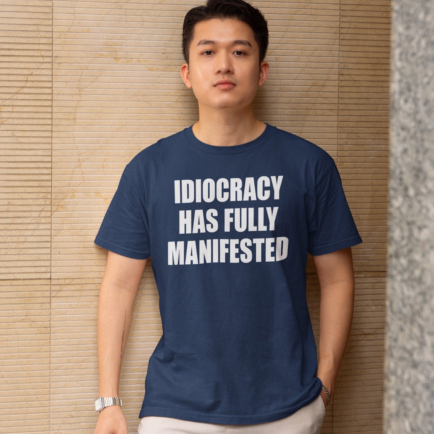 Young man wearing the Idiocracy T-shirt with bold white text, “IDIOCRACY HAS FULLY MANIFESTED,” standing against a tiled wall, embodies a statement piece for the politically aware.