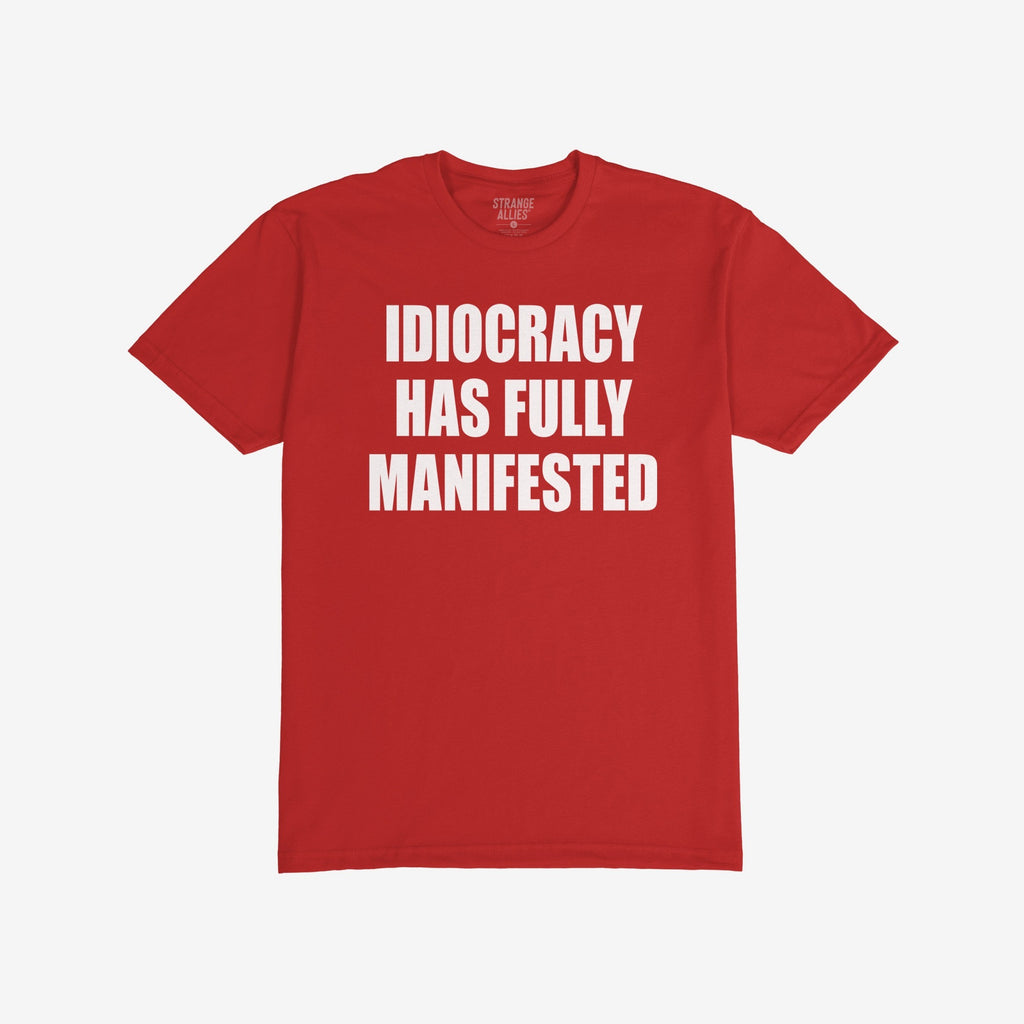 Idiocracy T-shirt with bold white text: IDIOCRACY HAS FULLY MANIFESTED. Brand label STRANGE ALLIES visible in collar. A statement piece for those embracing blunt truth and political commentary.