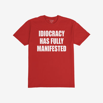 Idiocracy T-shirt with bold white text: IDIOCRACY HAS FULLY MANIFESTED. Brand label STRANGE ALLIES visible in collar. A statement piece for those embracing blunt truth and political commentary.