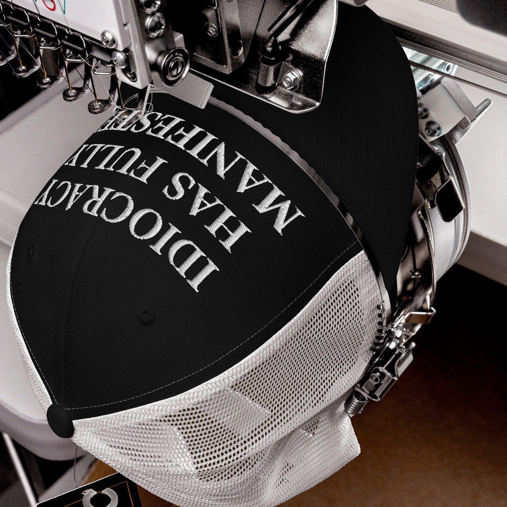 Black-and-white Idiocracy Hat in embroidery machine; reads “IDIOCRACY HAS FULLY MANIFESTED” in serif type, reflecting bold social commentary. Needles and thread spools in action, defining its creation process.