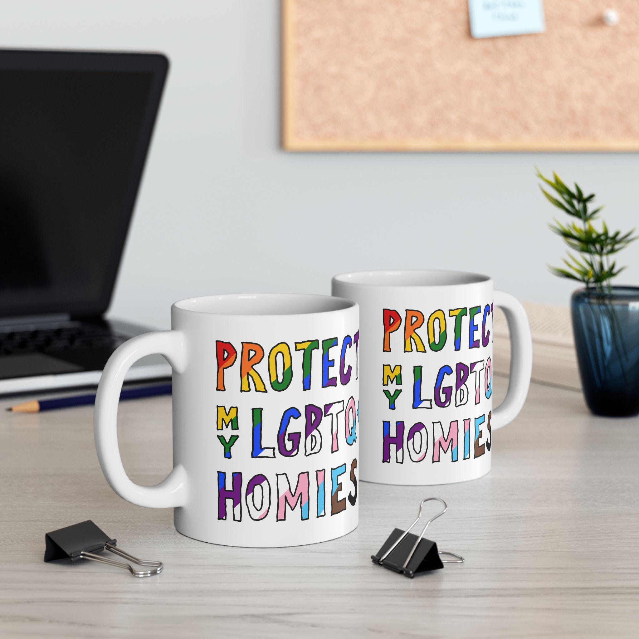 Protect My LGBTQ+ Homies Mug on a wood desk beside a laptop and stationery, showcasing bold, rainbow-colored lettering that promotes LGBTQ+ support, ideal for everyday use and gifting.
