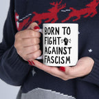 Person holding the Born To Fight Against Fascism Mug, featuring bold black lettering and a raised fist graphic, set against a dark knit sweater, embodying resistance and progressive ideals.