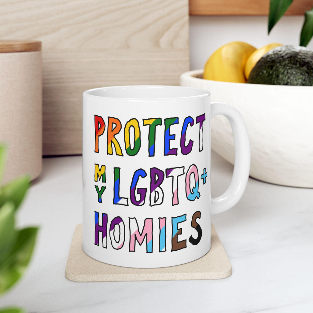 Protect My LGBTQ+ Homies Mug on coaster, featuring vibrant, hand-drawn lettering. Ideal for allies and community members, this ceramic mug embodies everyday support and solidarity. Dishwasher-safe and available in two sizes.