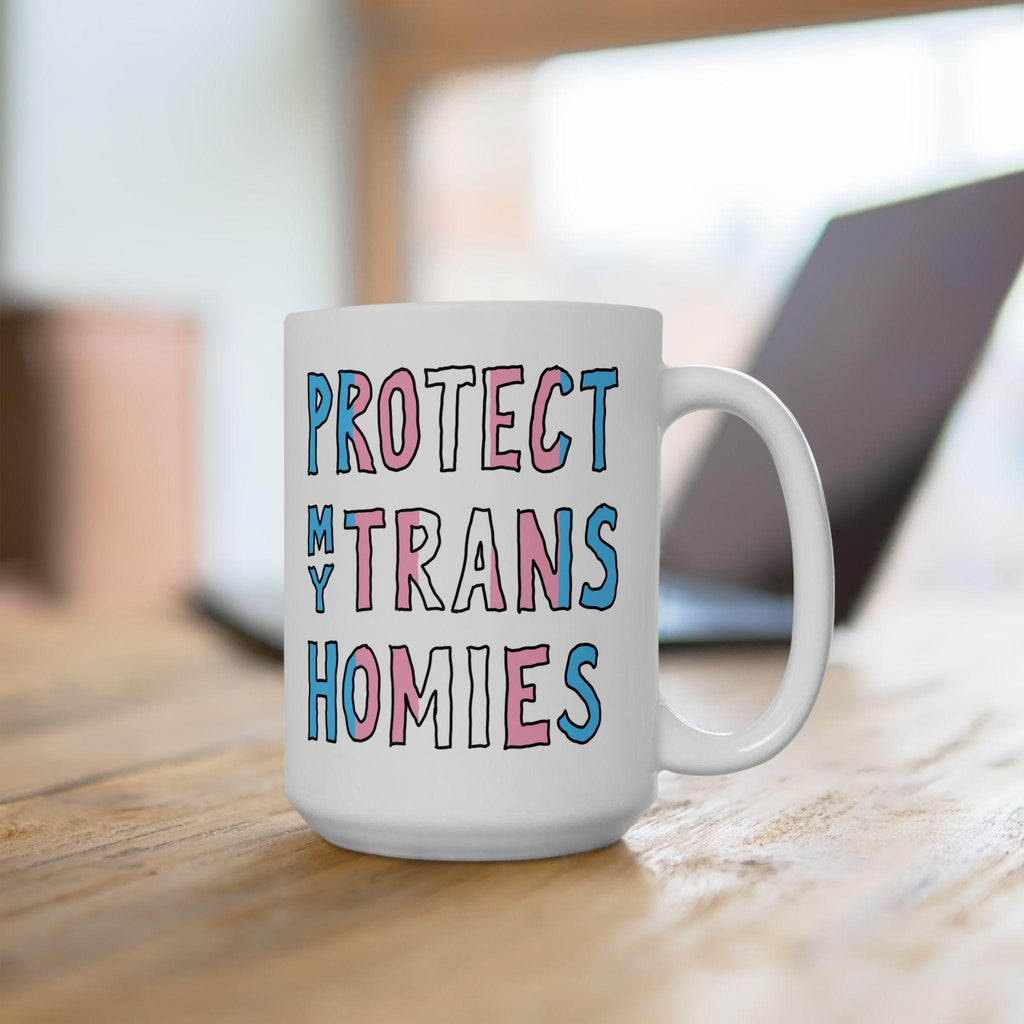 White ceramic Protect My Trans Homies Mug on a wooden table, featuring bold block letters filled with transgender pride colors, promoting equality and respect. Background shows a blurred laptop outline.