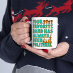Person in a dark sweater holds the Your Favorite Band Has Always Been Political mug featuring bold turquoise and orange text with raised fists and flames, embodying rebellion and activism.