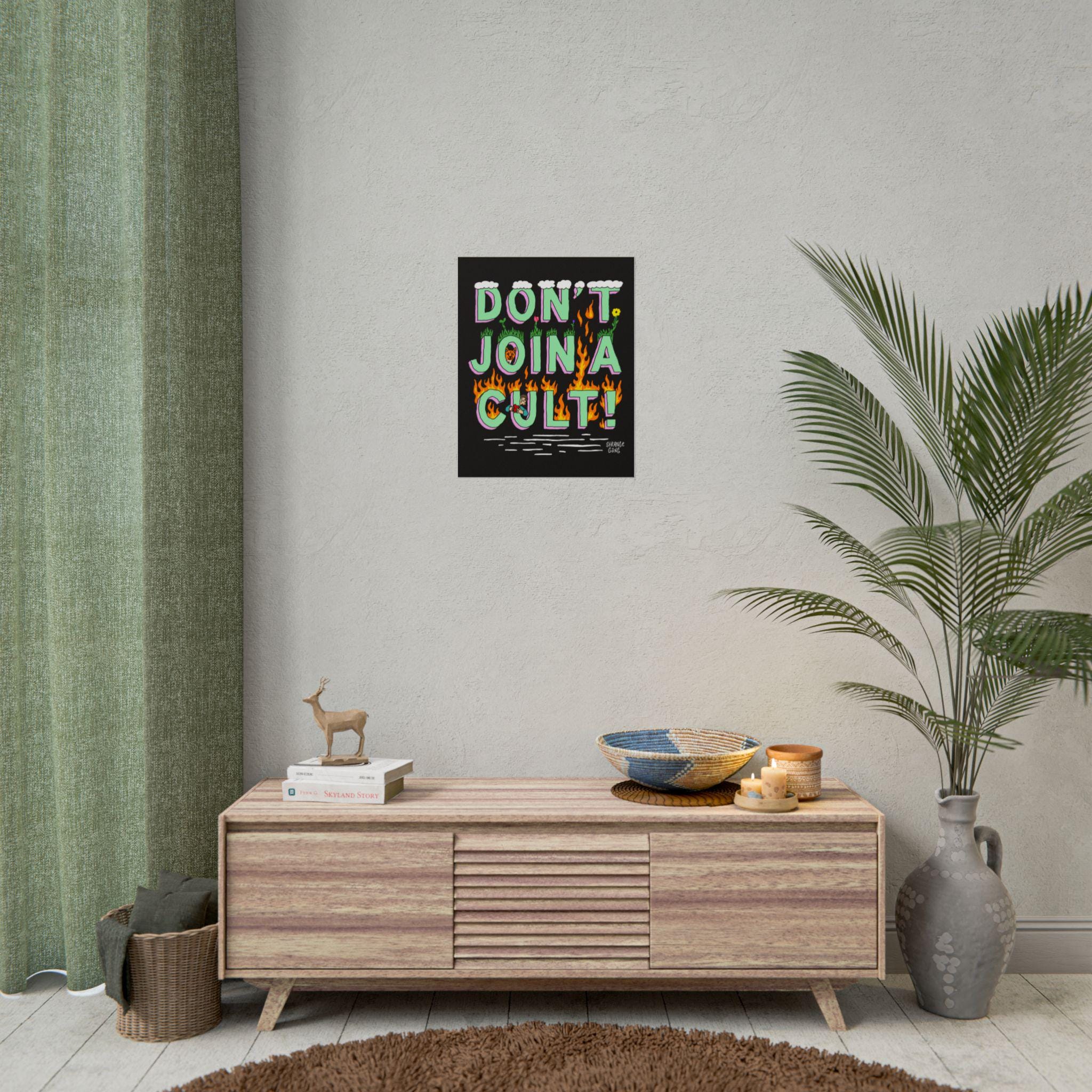 Don't Join A Cult Poster with fiery lettering on a light wall, above a wooden sideboard with books and a deer figurine, surrounded by decor like a woven basket and lit candles.