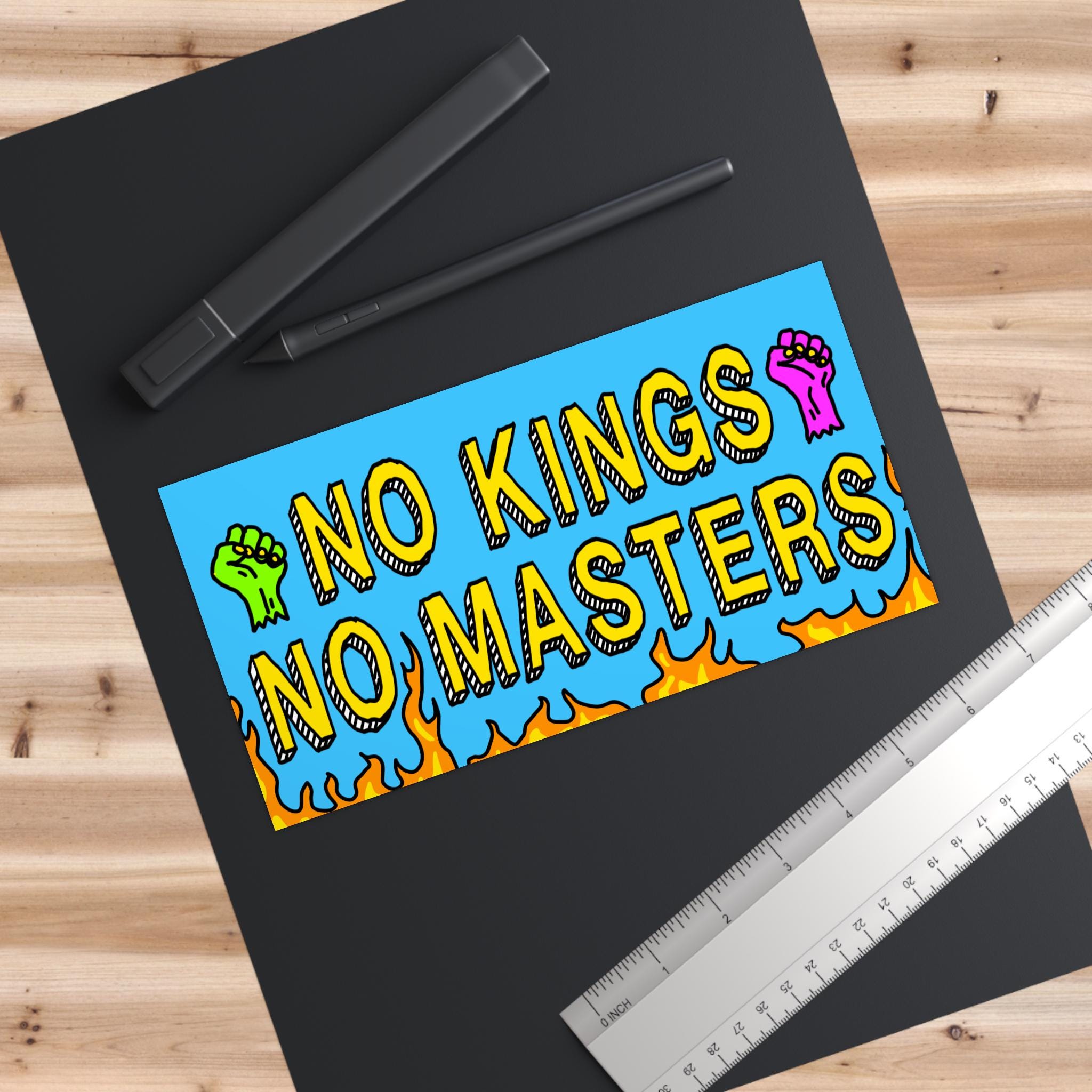 No Kings No Masters sticker on a desk, featuring bold yellow text and raised-fist icons, symbolizing resistance. Set against a blue background with orange-yellow flames, embodying defiance and activism.