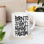 Born To Fight Against Fascism Graffiti Mug on cork coaster with bold, hand-lettered text, embodying resistance. Background includes a wooden cutting board, fruit bowl, and leafy plants, aligning with the store's activist ethos.