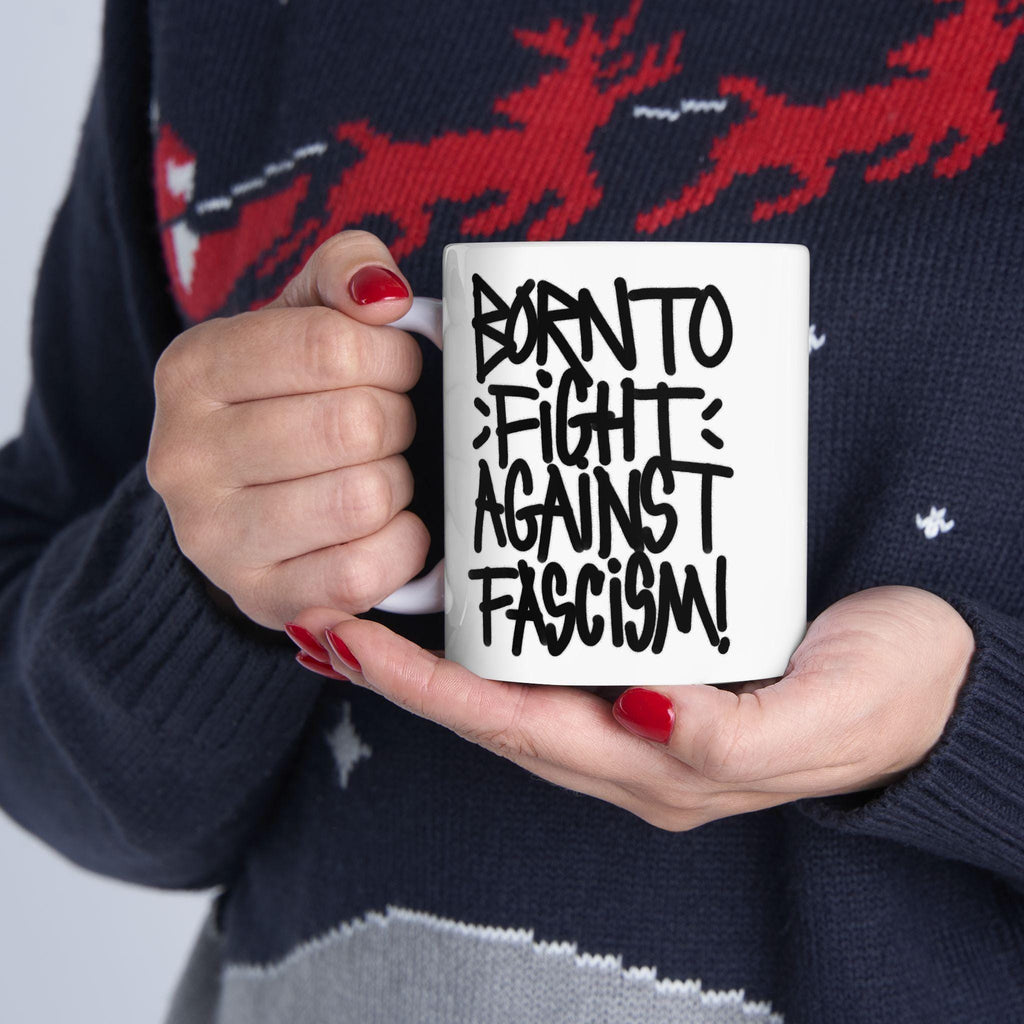 Born To Fight Against Fascism Graffiti Mug held by hands with red nails, displaying bold, black graffiti slogan. Person wears dark sweater with red reindeer motif, embodying a spirit of resistance.