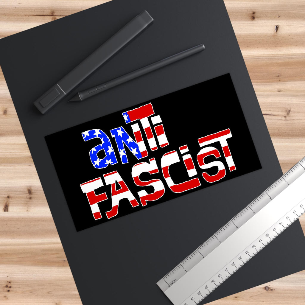 Top-down view of the Anti Fascist USA Sticker & Magnet on a black sketchpad, featuring a stars-and-stripes motif, alongside two digital-stylus pens and a metal ruler.