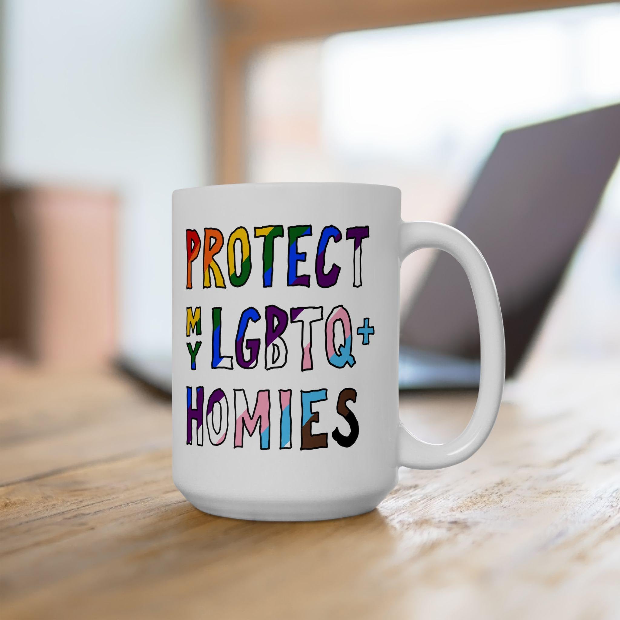 Protect My LGBTQ+ Homies Mug on desk, featuring colorful hand-drawn text supporting LGBTQ+ rights. Background includes blurred laptop and office elements, emphasizing daily solidarity and vibrant advocacy.
