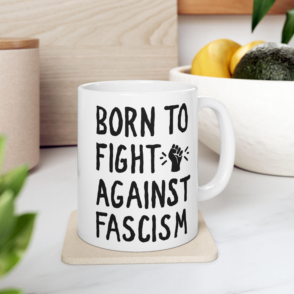 White ceramic Born To Fight Against Fascism mug with bold black lettering and a raised-fist icon, sitting on a coaster. Perfect for progressive coffee lovers advocating for equality and resistance.