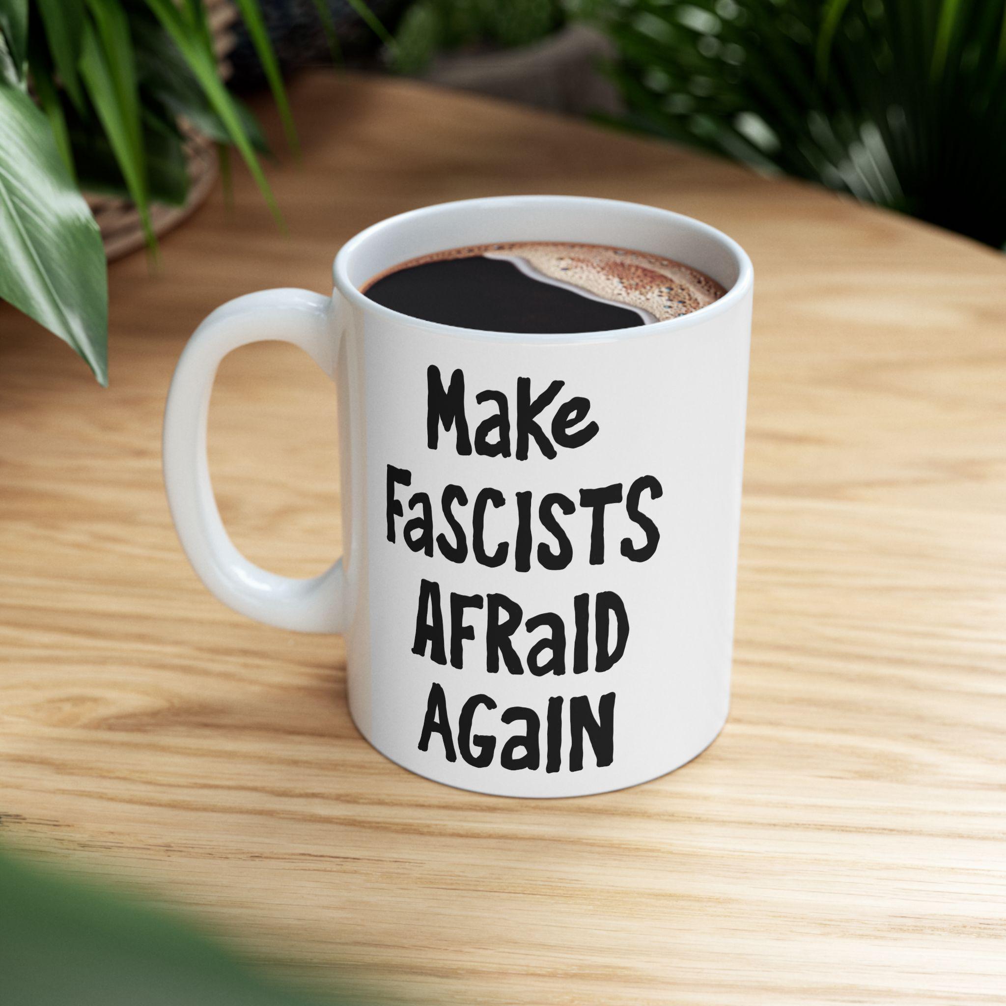 Make Fascists Afraid Again Mug on a wooden tabletop, featuring bold hand-lettered slogan. Filled with black coffee, this ceramic mug embodies resistance and activism, designed to inspire courage and defiance.