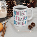 Protect My Trans Homies Mug with hand-drawn text sits on a light-gray cloth, surrounded by pine cones, a candle, cinnamon sticks, an orange, and twinkling lights, symbolizing warmth and support for equality.