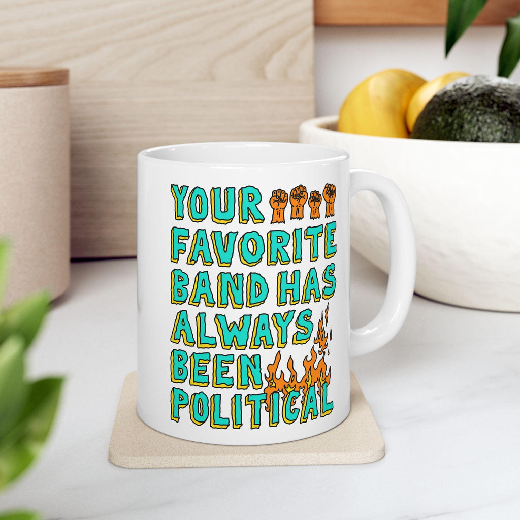 White ceramic Your Favorite Band Has Always Been Political mug on cork coaster, featuring vibrant hand-drawn text with raised fists and flames, aligning with rebellious music and social justice themes.