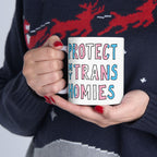 Person holding a white ceramic Protect My Trans Homies Mug, featuring bold pink, white, and blue block letters. The mug symbolizes support for trans lives, echoing themes of equality and justice.