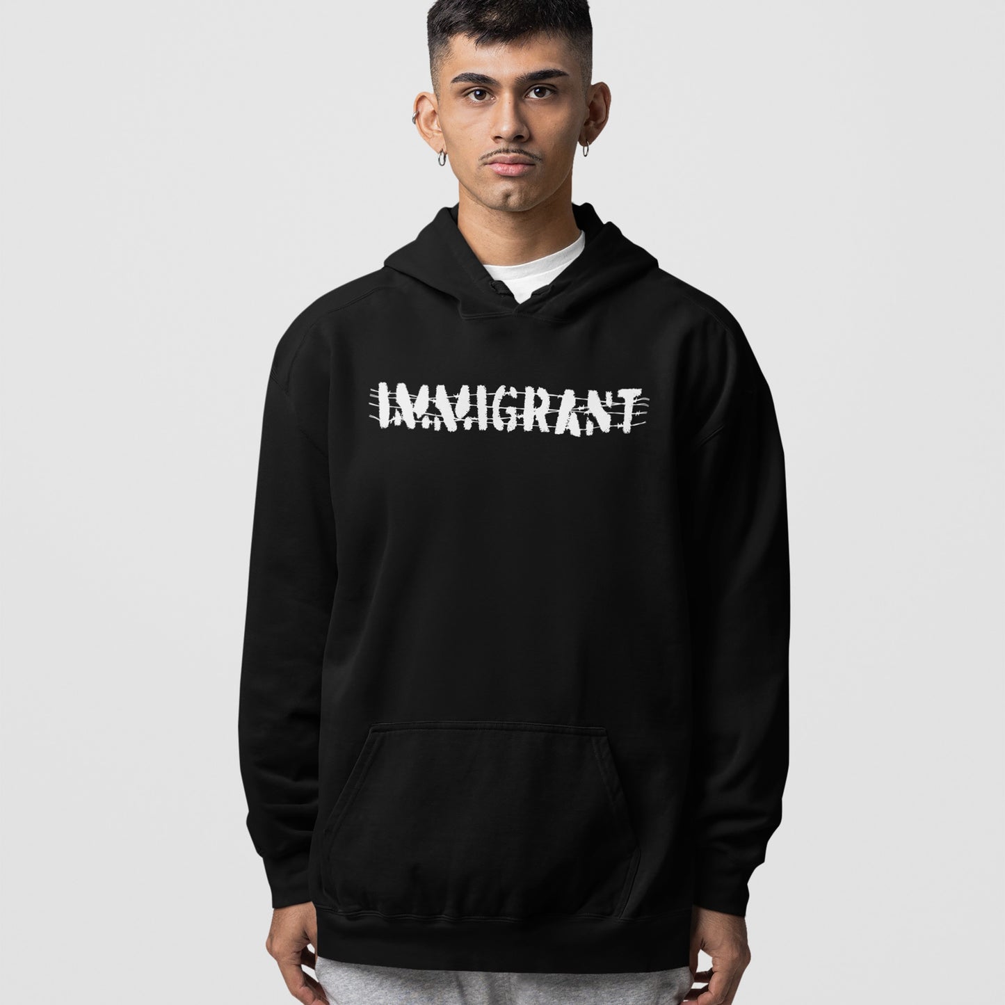 Young man wears the Immigrant Shirt hoodie, featuring bold white lettering crossed out with scribbles, symbolizing resilience and unity.