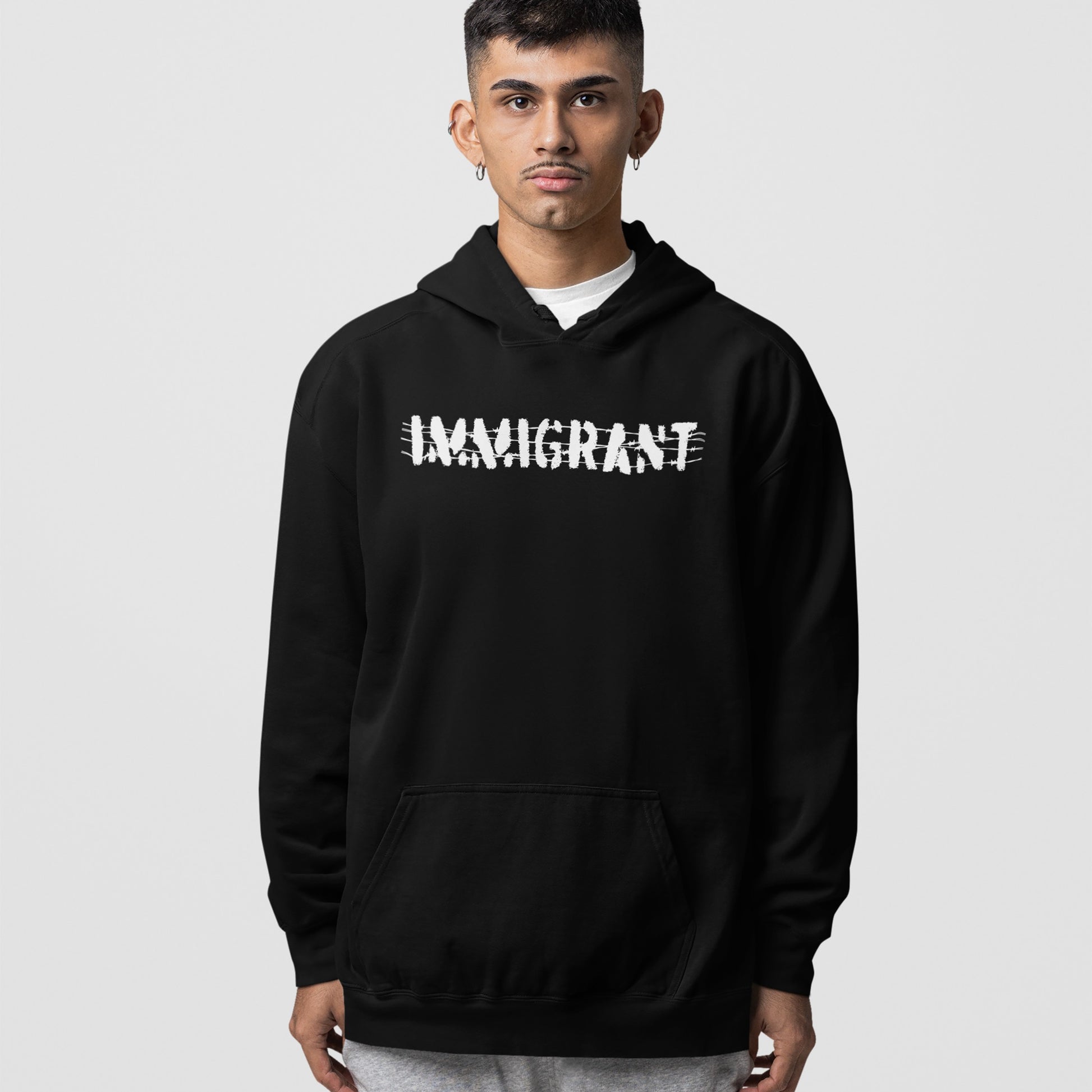 Young man wears the Immigrant Shirt hoodie, featuring bold white lettering crossed out with scribbles, symbolizing resilience and unity.