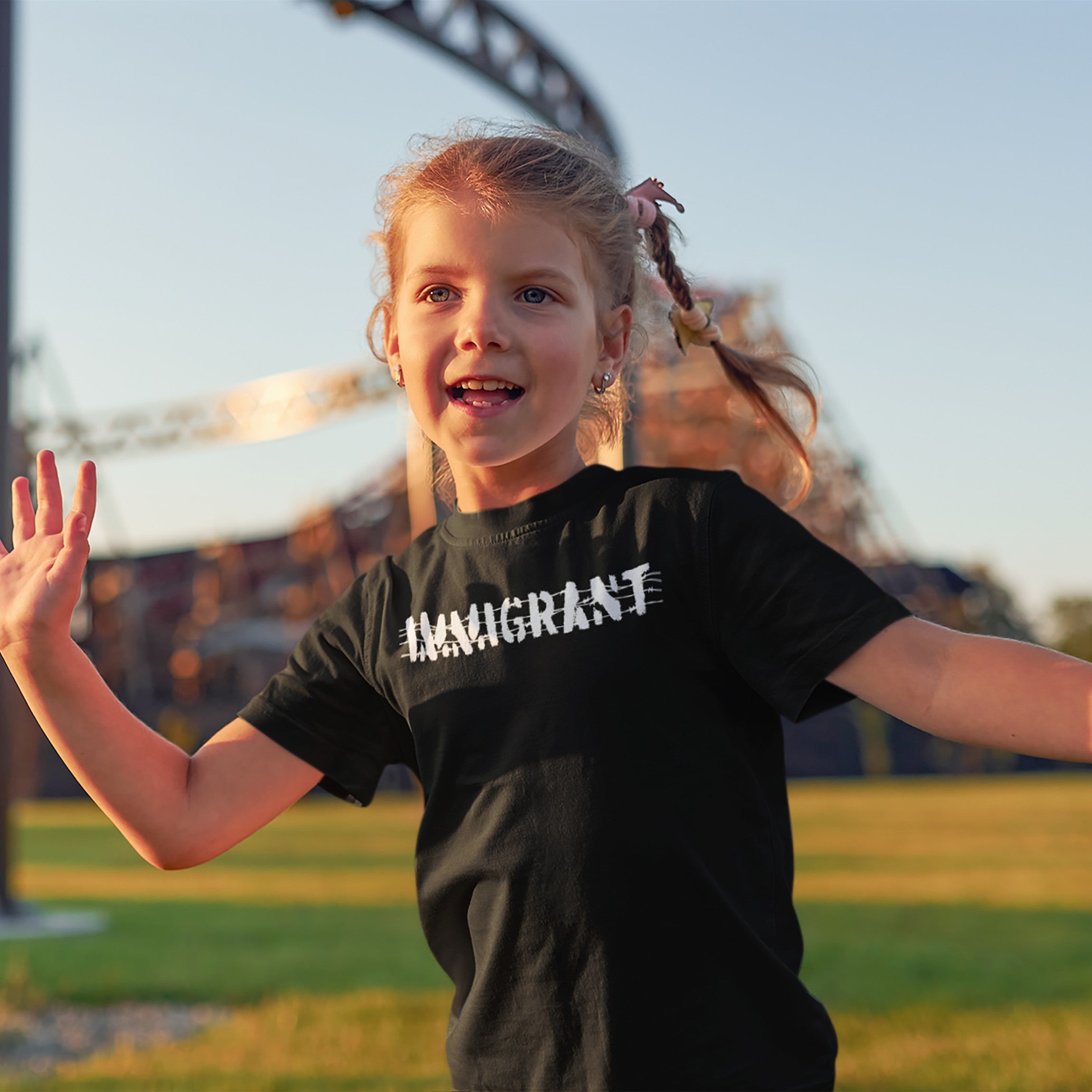 Young girl at a fairground wears the Immigrant T-shirt, featuring bold stencil lettering and barbed wire design, symbolizing resilience and solidarity with immigrant communities. She smiles, embodying activism and justice.