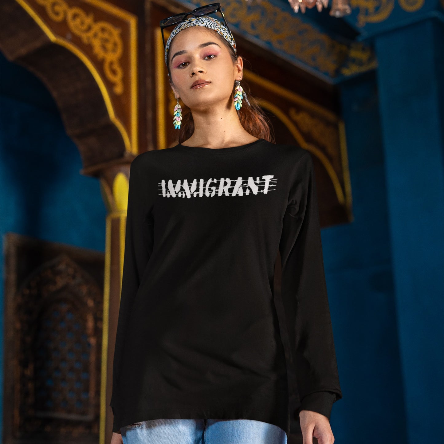 Young woman in a black long-sleeve Immigrant Shirt with IMMIGRANT crossed out, light-wash jeans, and a patterned headband, embodying resilience and unity against an ornately decorated background.