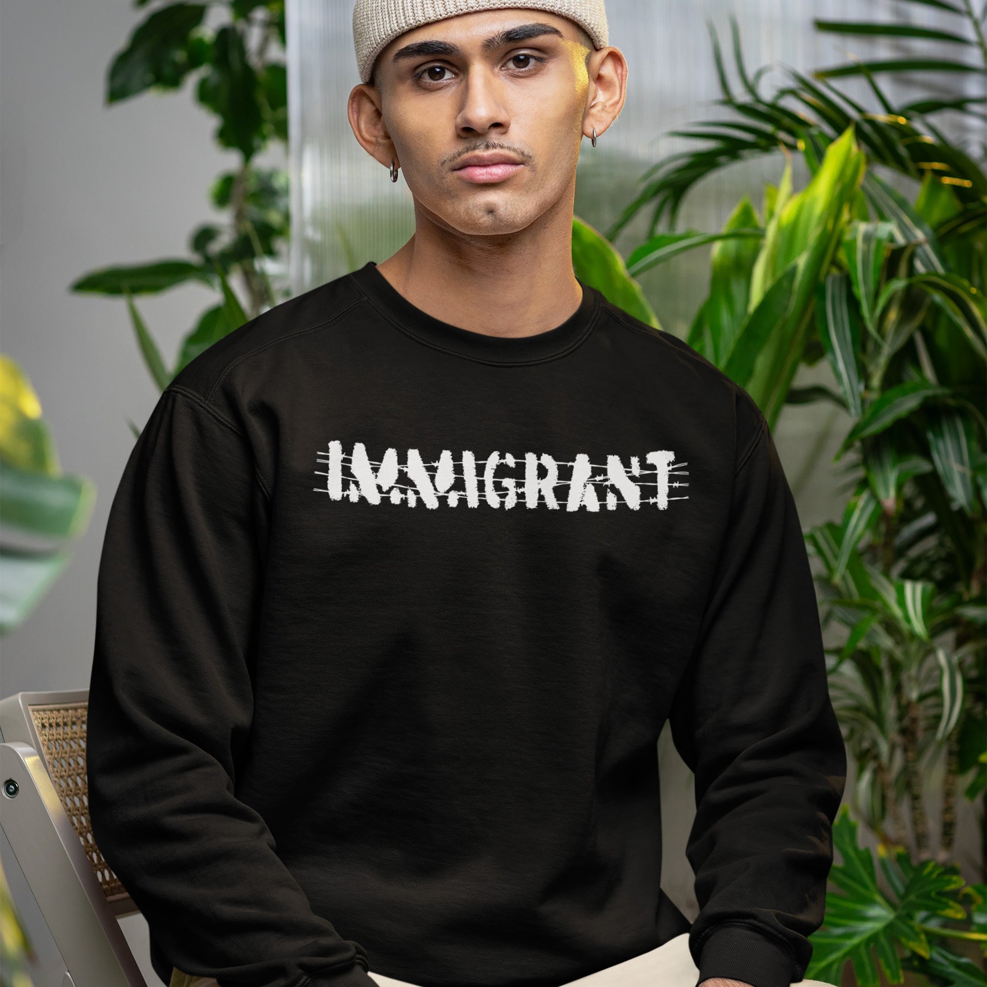 Young person wearing an IMMIGRANT sweatshirt with a scratched-out effect, seated in a plant-filled space, embodies resilience and solidarity, reflecting the bold statement of the Immigrant Shirt collection.