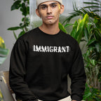 Young person in plant-filled space wearing the Immigrant Hoodie & Sweatshirt, featuring the word “IMMIGRANT” crossed out with brush-strokes, symbolizing resistance and solidarity with immigrant communities.