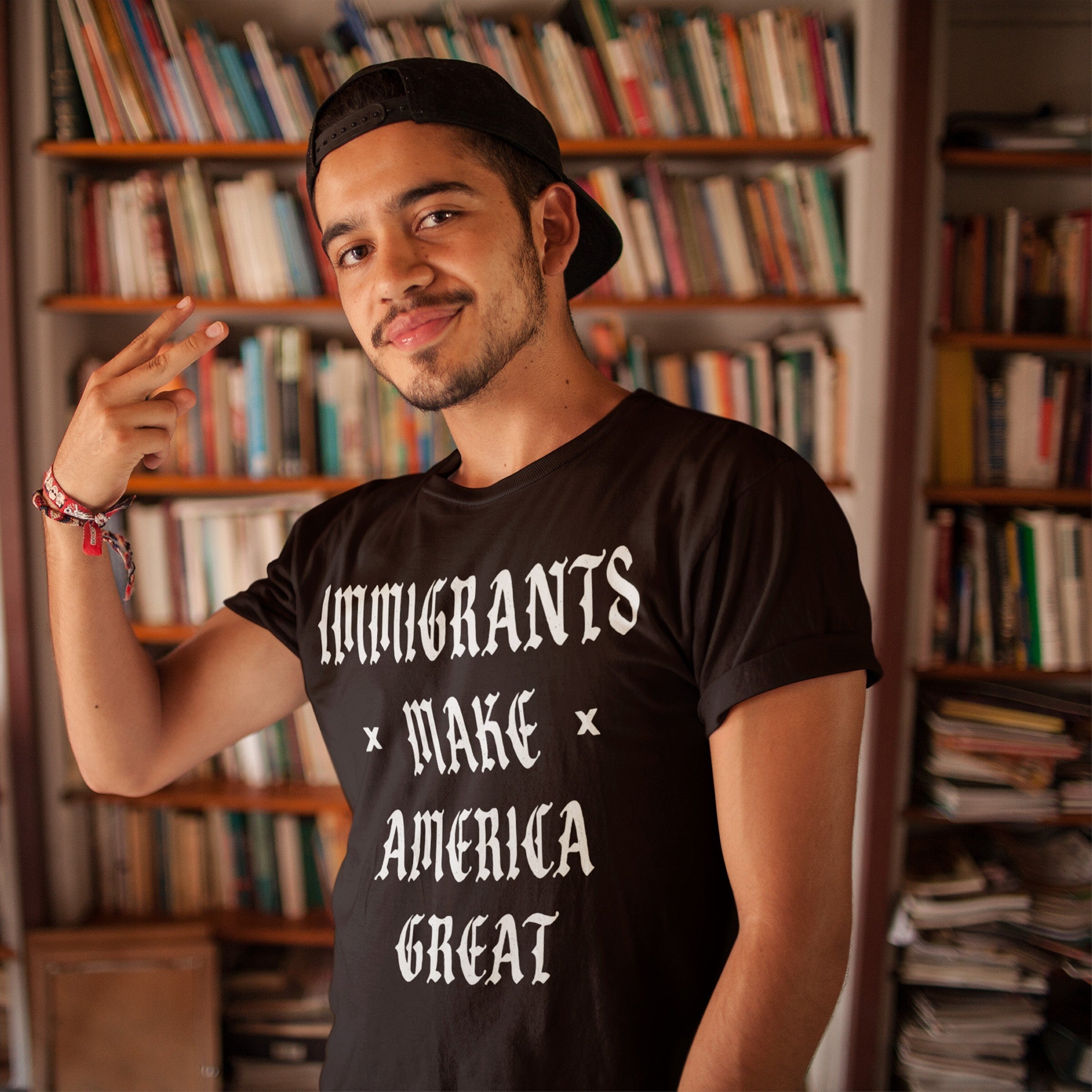 Young man in black T-shirt with “IMMIGRANTS MAKE AMERICA GREAT” slogan, standing before a bookshelf, smiling and flashing a peace sign. This protest-ready shirt supports immigrant rights and justice.
