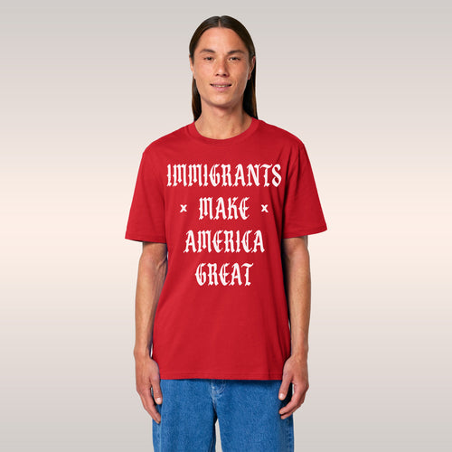 Young man in black T-shirt with “IMMIGRANTS MAKE AMERICA GREAT” slogan, standing before a bookshelf, smiling and flashing