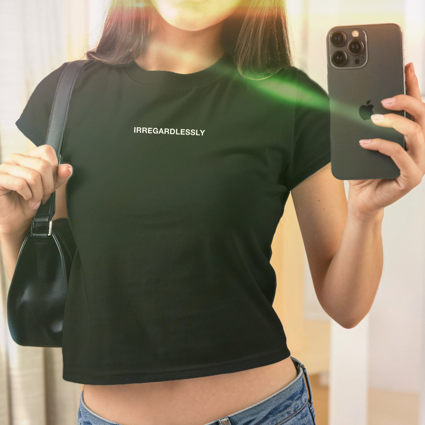 Mirror selfie featuring the Irregardlessly Women's Baby Tee, a black crop top with white lettering. A black shoulder bag and iPhone are visible. The tee reflects bold, rebellious style.