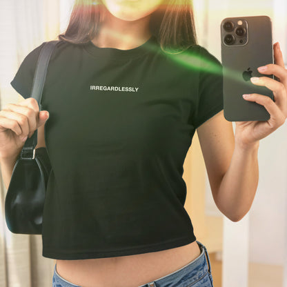 Mirror selfie featuring the Irregardlessly Women's Baby Tee, a black crop top with white lettering. A black shoulder bag and iPhone are visible. The tee reflects bold, rebellious style.