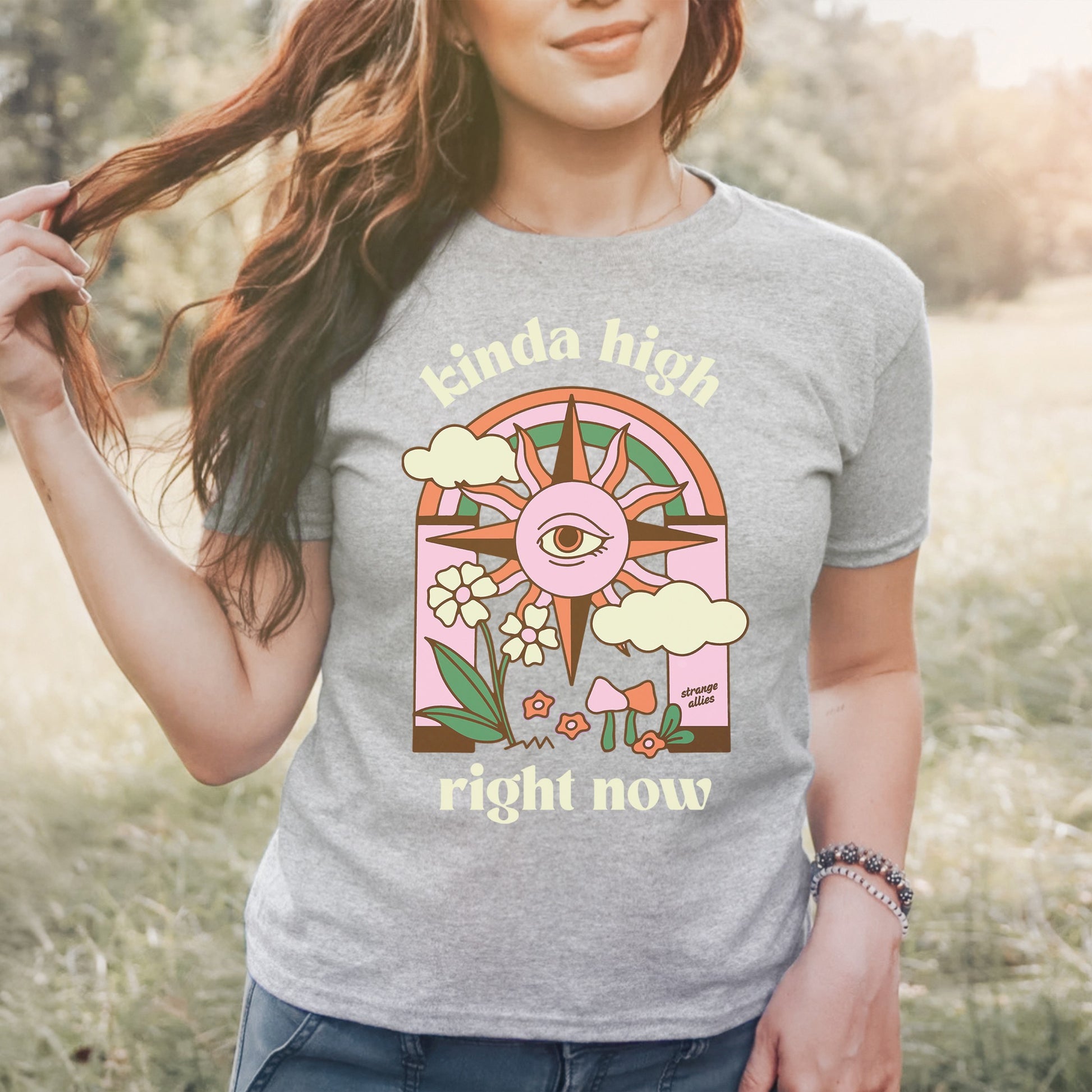 Woman outdoors wearing Kinda High Women's Baby Tee, featuring a colorful retro graphic with an all-seeing eye, sunburst, rainbow, clouds, flowers, and mushrooms, expressing a carefree, whimsical vibe.