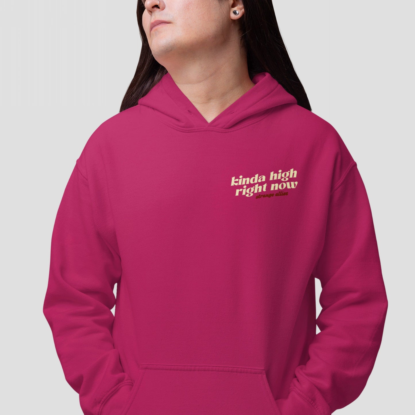 Person in a magenta Kinda High Hoodie & Sweatshirt with cream text, featuring kinda high right now and strange allies. Long dark hair, nose ring, light-gray background.