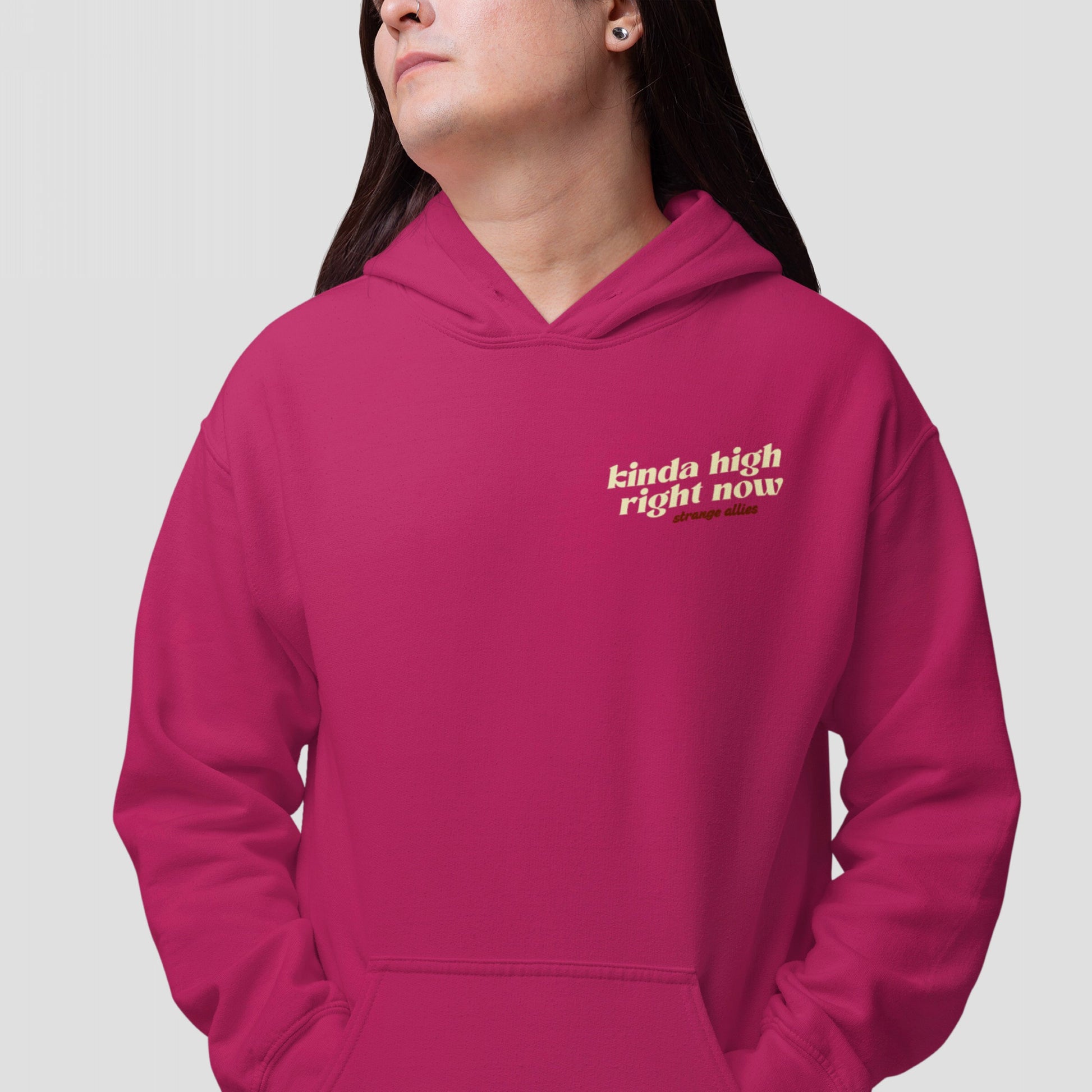 Person in a magenta Kinda High Hoodie & Sweatshirt with cream text, featuring kinda high right now and strange allies. Long dark hair, nose ring, light-gray background.