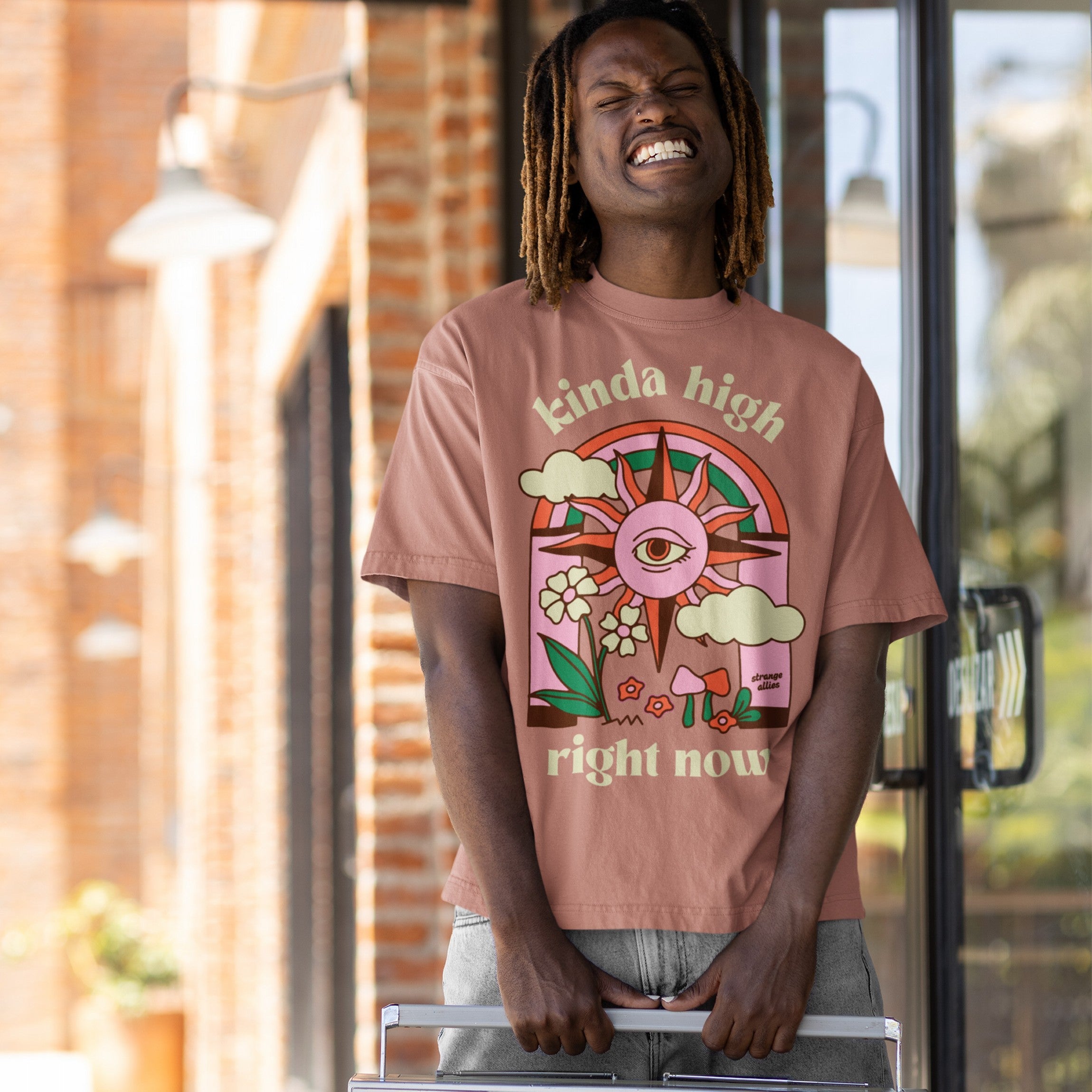Young man in a playful grin wearing the Kinda High T-shirt, featuring retro-psychedelic graphics with clouds, mushrooms, and a sunburst eye, symbolizing a mellow, introspective vibe.
