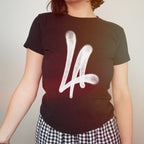 Person wearing the LA Handstyle Women's Baby Tee, featuring a bold, white LA graffiti graphic on a black background, paired with black-and-white checkered bottoms, capturing a retro Y2K vibe.