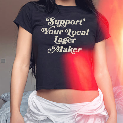 Support Your Local Lager Maker Women's Baby Tee displayed on a torso, showcasing its cropped fit and bold slogan. Perfect for expressing support for local brewers with style and attitude.