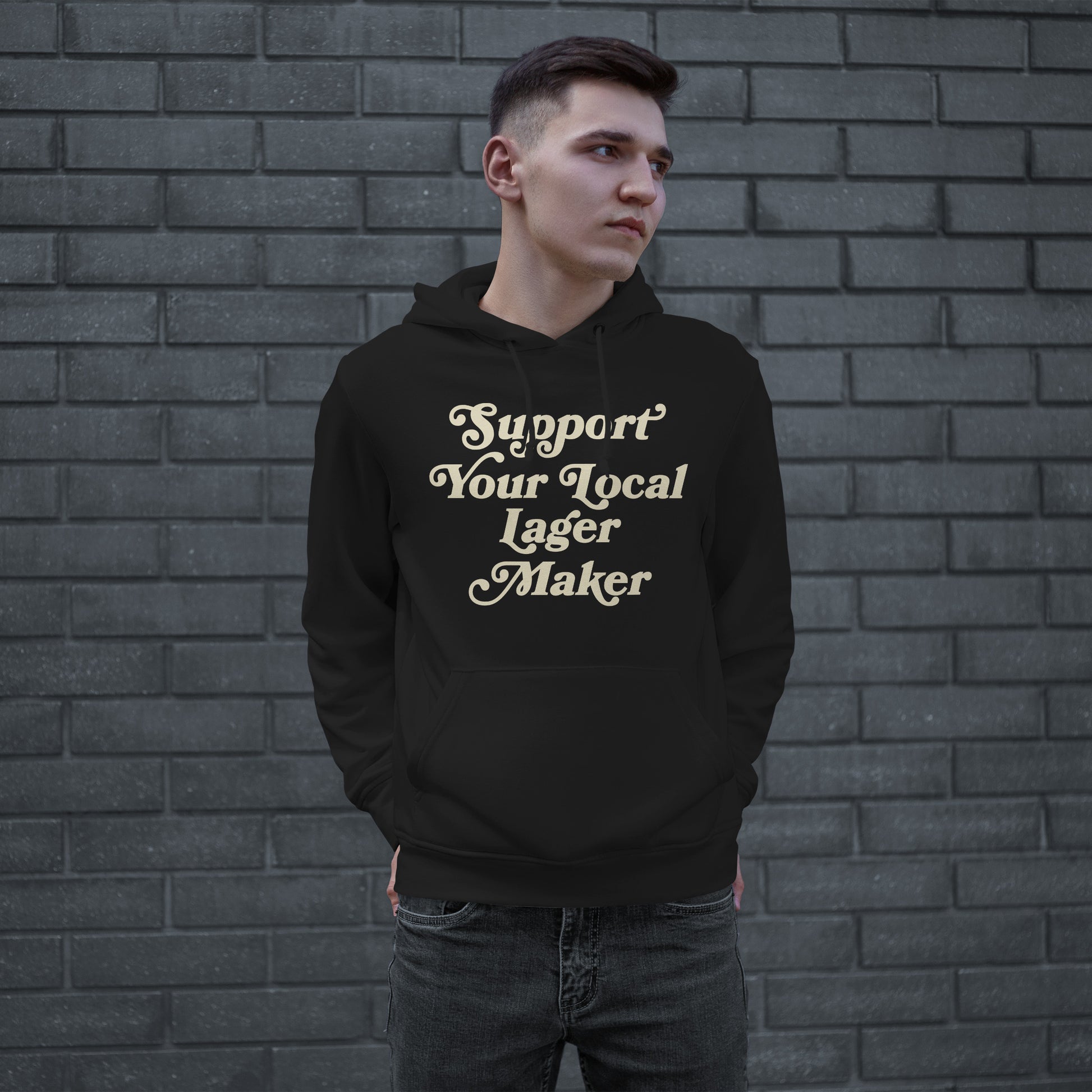 Young man in black Support Your Local Lager Maker hoodie, featuring cream vintage script, stands against a brick wall, embodying the ethos of supporting local craft beer and community pride.
